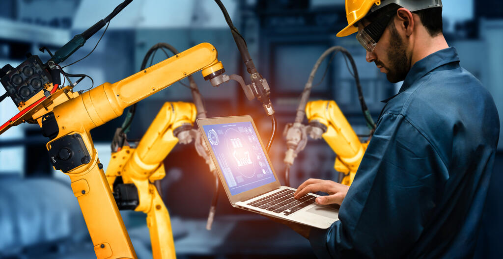 A worker working on a laptop, surrounded by helper industrial robots