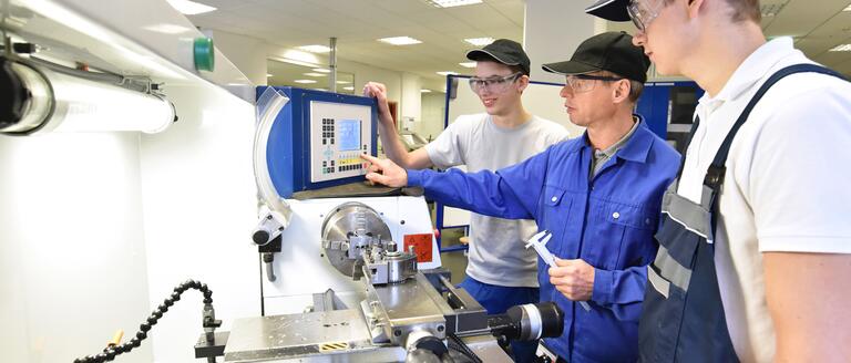 3 men wearing safety glasses clustered around a machine, working together collaboratively to solve problems.