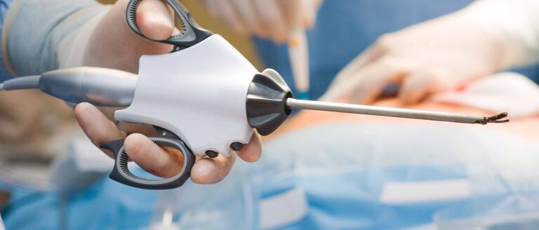 A hand holding an ultrasonic surgical tool with an operation occurring in the background