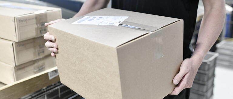 A man in a dark t-shirt holding a cardboard box next to a conveyor belt
